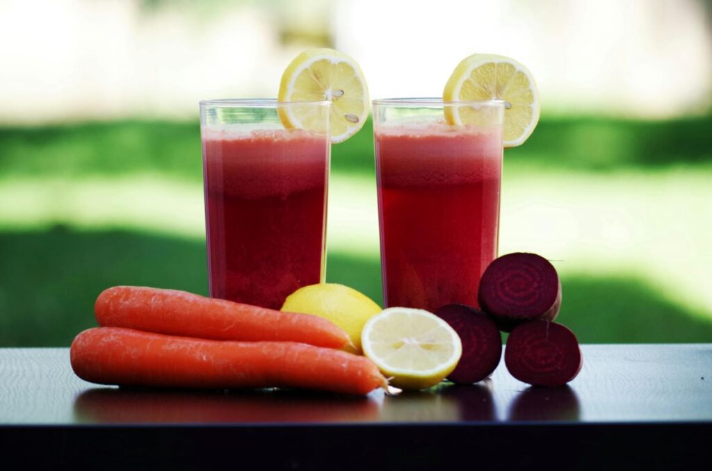 Two glasses of fresh red juice with ingredients like carrots and beets on a table.
